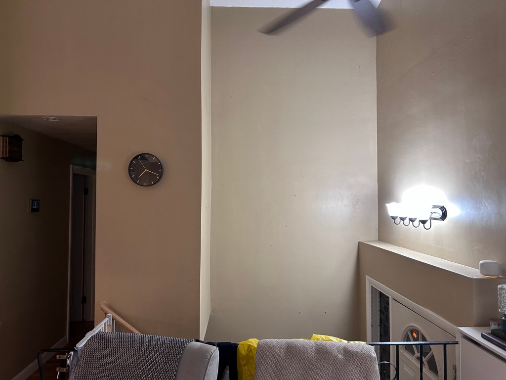 An interior view of a light-colored hallway featuring a wall clock, a ceiling fan, and a light fixture illuminating a corner.