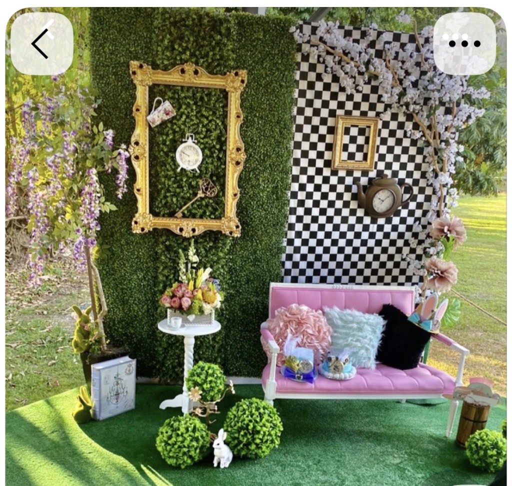 A whimsical outdoor scene featuring a pink vintage-style couch adorned with decorative pillows, surrounded by artificial greenery. In the background, a green foliage wall is paired with a black and white checkered pattern. Decorative elements include flower arrangements, framed artworks, a clock, and playful accents like stuffed bunnies and topiary balls.