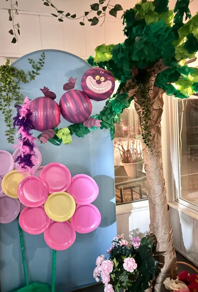 A whimsical display featuring a tree made of paper, colorful paper flowers, and painted spheres resembling the Cheshire Cat from Alice in Wonderland, set against a blue backdrop.