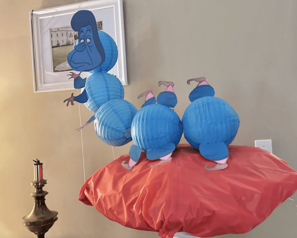 Colorful blue caterpillar character made from paper lanterns, sitting on a red surface, with a whimsical facial expression and cartoonish features.