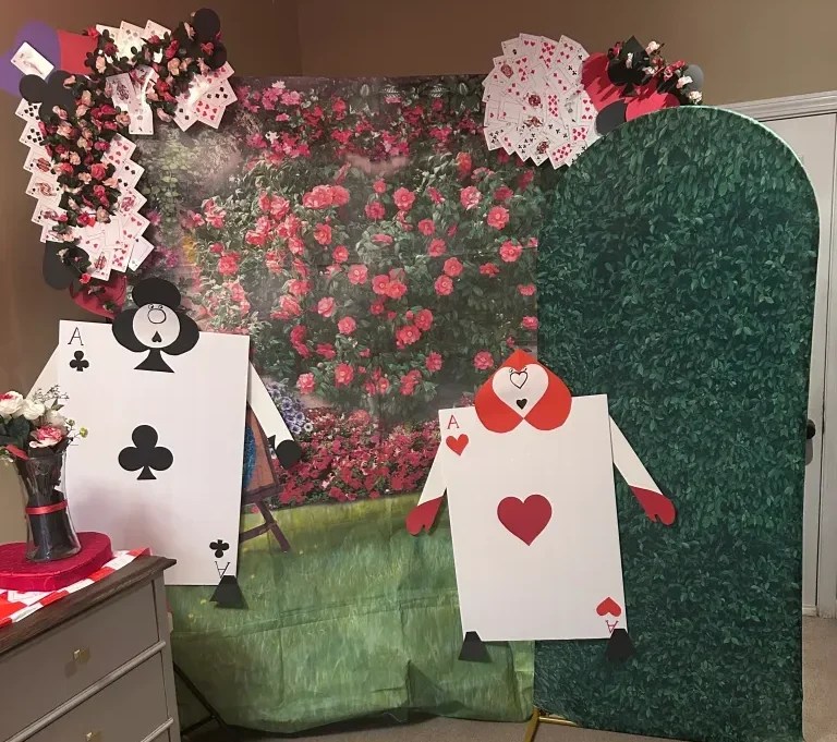 A whimsical backdrop featuring a garden of red flowers, with cutouts of playing cards: a black spade and a red heart. Decorative elements include paper playing cards and a floral arrangement in a black vase.