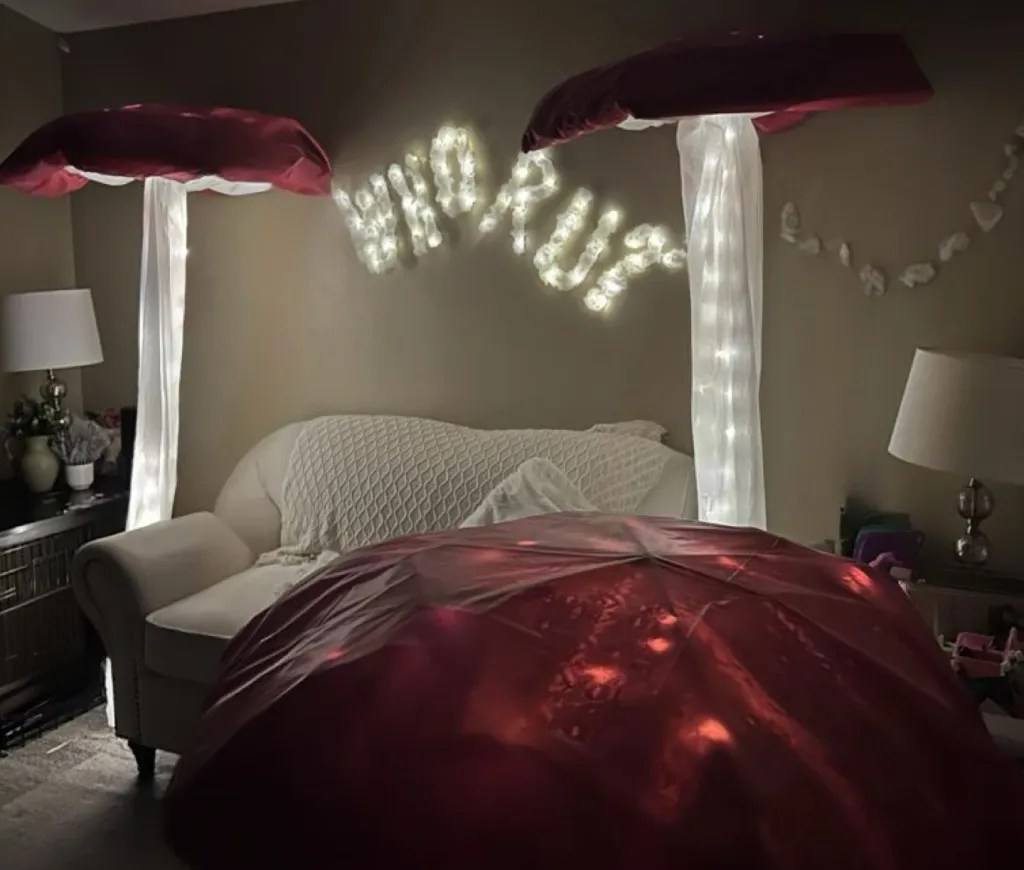 A cozy living room featuring a white couch with a knit blanket, illuminated by large red mushroom-shaped decorations with white drapes and string lights spelling 'WHO RU?' on the wall.
