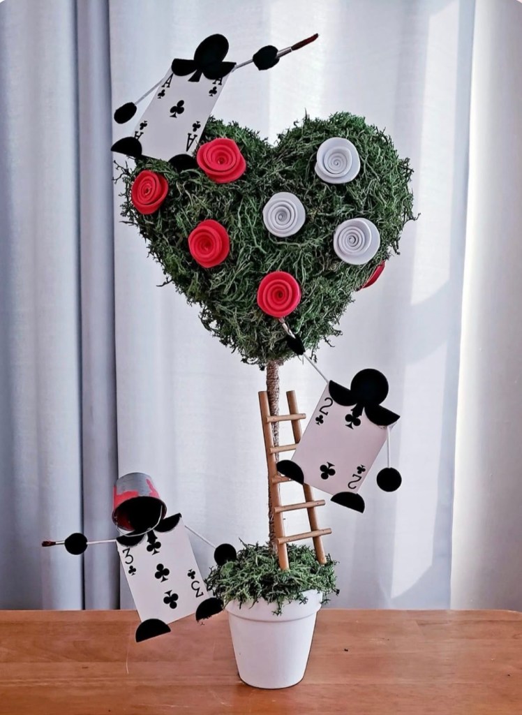 A decorative heart-shaped topiary made of green moss, adorned with red and white rolled paper flowers, and featuring playful elements like card-themed figures and a small wooden ladder.