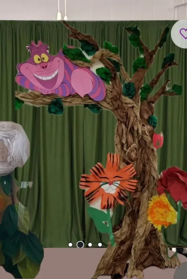 A whimsical tree with colorful flowers and a smiling Cheshire Cat figure lounging on a branch, displayed in front of a green curtain.
