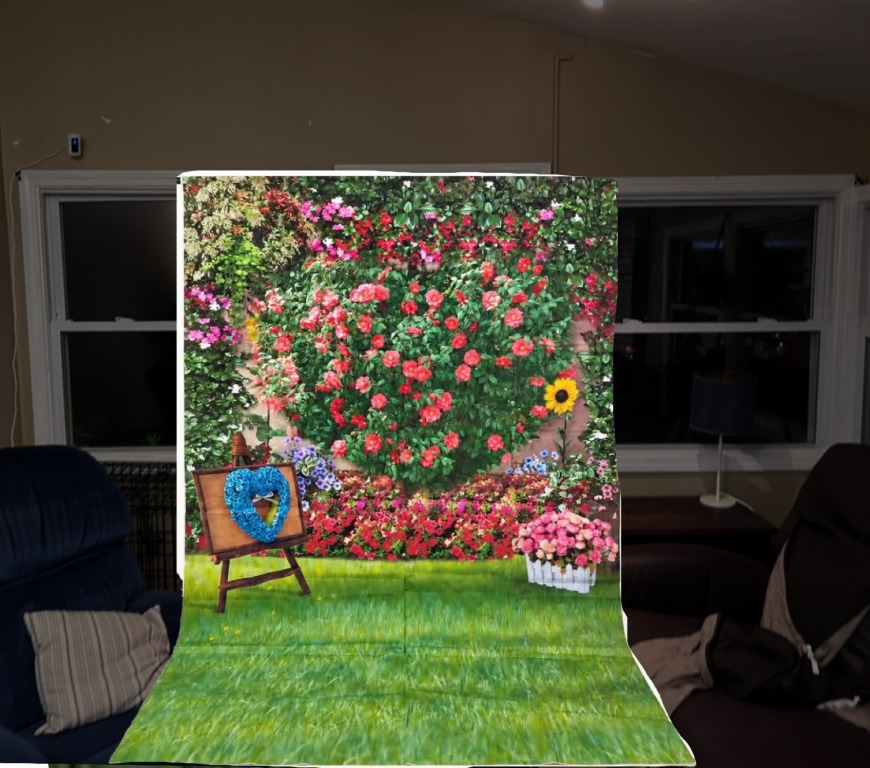 A decorative backdrop featuring a vibrant wall of blooming flowers in various colors, with a small wooden easel displaying a blue heart design in front of it. The scene is set on a green, grassy floor.